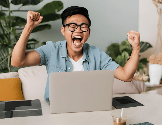 An Indonesian man cheers triumphantly at his laptop, arms up in victory and delight.